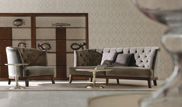 This picture shows an elegant and luxurious white marble bathroom with a large, rectangular freestanding bathtub. In the background is a tall window with drapery, as well as a wooden vanity with his and her sinks. There is a beautiful chandelier hanging from the ceiling and the floor is tiled with gold accents.