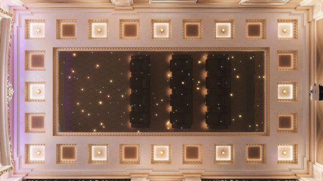 The picture shows a beautifully designed grand staircase made up of marble. The stairs are curved and the railing is golden with intricate detailing. The walls are painted white and the stairs are topped with a large, ornate chandelier. The flooring is a light-colored marble giving the room a luxurious atmosphere.