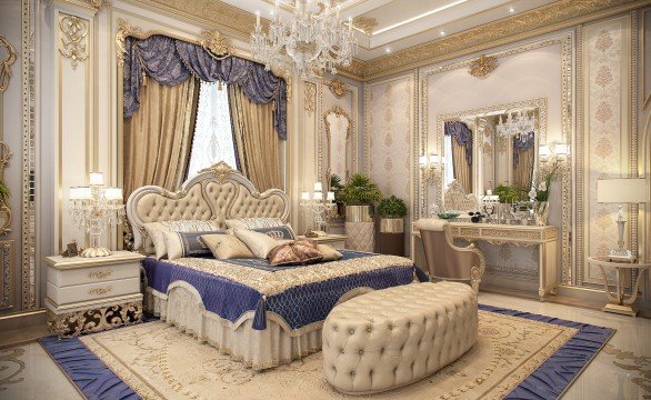 This picture shows a luxurious bedroom area in an upscale home. The room is furnished with a four-poster bed adorned with a tufted headboard, white bedding, and large pillows. It also features two bedside tables with lamps on top and a white tufted leather chaise lounge at the foot of the bed. The walls are finished in a silvery wallpaper and furnished with wall art, while the ceiling has an elaborate chandelier. The hardwood floors are covered by a large area rug.