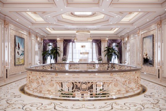 The picture shows a grand entrance hall to an upscale home or estate. It features white and gold accents, with a double staircase leading up to a balconied landing. The walls are adorned with large black floral designs, as well as intricate wrought iron detailing. A large, ornate chandelier hangs from the ceiling. On either side of the staircase are two marble columns. The luxury and opulence of this entrance hall is further accentuated by the detailed pattern on the floor.