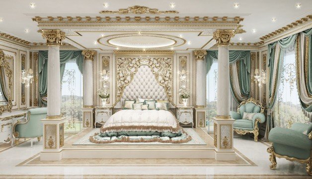 This picture shows a luxurious and grand living room with a statement wall feature. The walls are a light beige color, and there is a large white fireplace surrounded by a black marble trim. There is a luxurious white sectional sofa in the middle of the room with brown and cream pillows and a large gold coffee table in front of it. On either side of the seating area are ornate white armchairs with large tufted backrests. A large chandelier hangs from the ceiling, while two small gold side tables flank the sofa. Finally, an attractive framed painting hangs