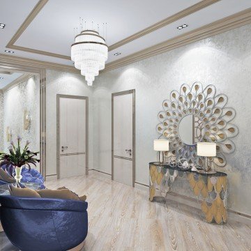 This picture shows an ornately designed living room decorated in a classic, yet modern style. The walls are painted a soft gray and are adorned with gold and silver accents. A white marble fireplace takes center stage and is flanked by two loungers upholstered in a rich velvet fabric. The floor is covered in a fashionable patterned area rug and there is a crystal chandelier hanging from the ceiling.