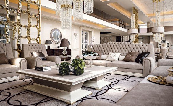 This picture shows a luxurious living room with a classic and modern design. The walls are painted in a light beige color and are decorated with beautiful wallpaper that features a cream-colored floral pattern. The furniture is a mix of brown, gray and white pieces that exude luxury. An impressive white chandelier hangs from the ceiling, providing a focal point for the room. A large rug sits in the center of the room, providing softness and warmth to the space.