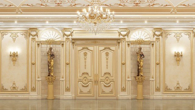 This picture shows a luxurious modern bedroom. The walls are decorated with an ornate gold and white patterned wallpaper, and the floor is covered in a light beige hardwood. A large four-poster bed is taking center stage, dressed with multiple layers of plush white linens and a tufted headboard. On each side of the bed, there stands a nightstand decorated with a lamp and several framed photographs. In the background, one can see two small sitting areas with a velvet couch and armchair and several decorative pillows.