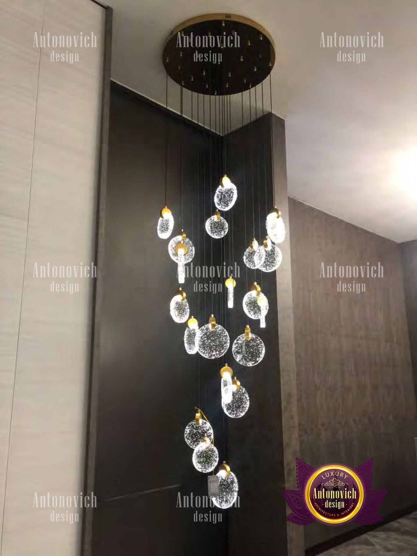 This picture shows an elegant, modern interior design. It features a curved staircase with metallic railings, a marble floor, and a combination of white and light grey walls. The atmosphere is completed by a modern chandelier and a large abstract painting hung on the wall.