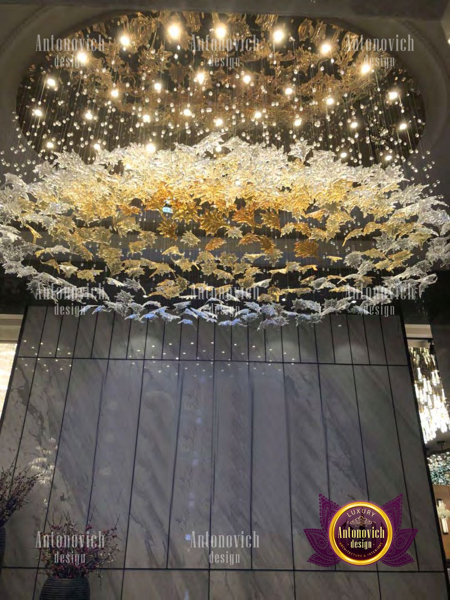 A luxurious and grand foyer featuring an intricate ceiling design, with a marble and wood staircase, elaborate wood wall panels, and ornate metal chandeliers.