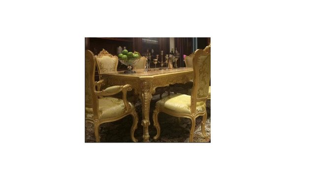 This picture depicts a luxurious living room in a home. It features a golden-colored sectional couch with classic-style armchairs and cushions, a marble-topped coffee table set on top of a white rug with an elaborate pattern, a stylish crystal chandelier overhead, and a decorative wooden sideboard in the corner. The walls are painted in a soft beige hue with ornate gold trim, while the windows are covered with semi-sheer drapes. A large mirror is mounted above the sideboard, accentuating the regal feel of the room.