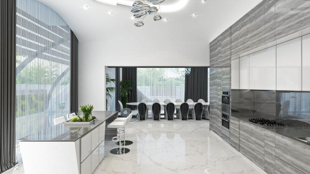 This picture shows a luxurious modern kitchen designed by Antonovich Design. The kitchen has a large island with seating, a granite countertop with a built-in sink, and a range with glass-fronted cabinets above it. The floor is tiled with a white-and-gray pattern and the walls are painted a light gray. The cabinets are white and feature stainless steel hardware. The kitchen also has recessed lighting and a large window that looks out onto a garden.
