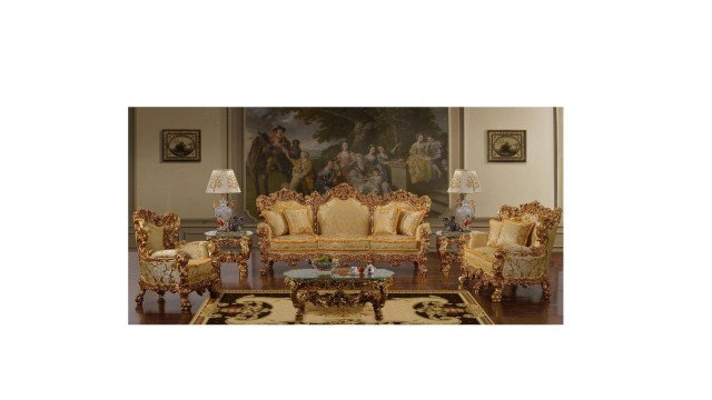 This picture shows a luxurious bedroom with a grand canopy bed and a delicate silk overlaid bedspread. The bed frame is made of dark wood and is adorned with intricate carvings and period-era details. The headboard includes a classic drapery with tassels and the walls are lined with mahogany paneling and beveled mirrors. Accented with works of art, a crystal chandelier hangs in the center of the room, illuminating a tufted chaise lounge and two ornate dove grey armchairs. Other luxurious pieces of furniture, such as a