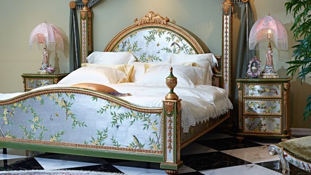 This picture shows a modern bedroom design featuring an ornately carved wooden bed frame with an upholstered headboard, sitting atop an intricately patterned rug. The room is furnished with neutral accent pieces, such as an armchair, a side table, and a sleek white nightstand. The walls are also given an elegant touch with ornate wallpaper and detailed cornices. Several mirrors add to the sophisticated look of the room and create a sense of brightness and airiness.