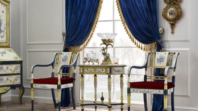 This picture shows an ornately decorated interior of a luxurious living room. It features a white grand piano against a white wall, which is accented by a gold and white fireplace mantel. The floor is covered by a white and gold patterned rug, and there are two plush white sofas with accent pillows. There are also two gold side tables and a few lamps for additional lighting. Finally, there is a large glass vase of vibrant pink and purple flowers as the centerpiece of the room.