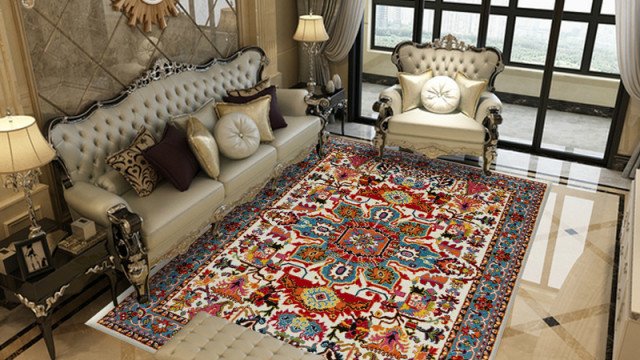 This picture shows a modern luxury living room interior with a dark gray velvet loveseat and armchair, two white accent chairs, a black round ottoman in the center of the room, and a unique black and white patterned rug. The color palette is mostly neutral with touches of black, white, and gray. There are several abstract art pieces hung on the walls, as well as a large wall-mounted TV.