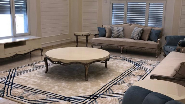 This picture shows a luxurious bedroom with a four-poster bed covered in white linen and surrounded by dark wood furniture. Gold accents add a hint of glamour to the space. The walls feature light gray wallpaper and ornate gold fixtures. A large window with sheer curtains allows natural light to stream into the room, while two white armchairs provide a cozy seating area.