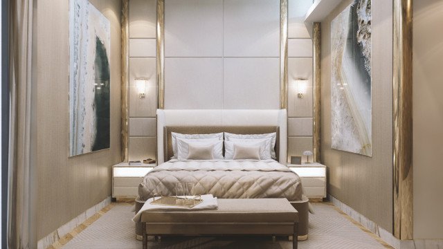 This picture shows a luxurious white and grey bedroom with a custom designed bed centered in the room. Two small, gold-framed night stands flank the sides of the bed, while two grey and white armchairs sit to either side. The walls are adorned with a pattern of intricate, gold and white designs, while a large glass chandelier hangs above the bed.