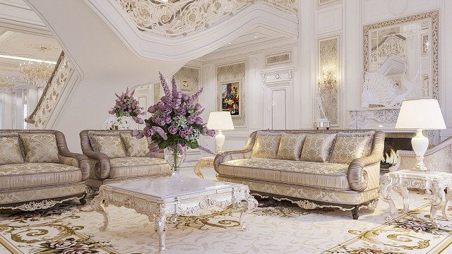 This picture displays a luxurious foyer in an elegant modern home. The walls and floor are finished in marble with intricate detailing. A grand staircase with an ornate railing leads to the upper floors, while a chandelier hangs from the ceiling. A set of double doors are open at the end of the foyer, leading out to a terrace area.