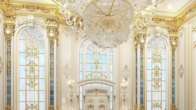 This picture shows a large, elaborately decorated foyer in an elegant home. The walls and ceiling are painted white, accented by crown molding and detailed trim works. The floor is covered with a luxurious marble pattern, and the centerpiece of the foyer is a grand staircase with intricate wrought iron railings and a curved marble banister. Several chandeliers hang from the ceiling, providing diffused light throughout the space.