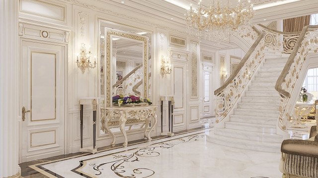 This picture shows a grand reception area in some kind of office, hotel, or commercial space. It has large crystal chandeliers, lush furniture, and gold accents throughout. The walls are ornately decorated with patterned wallpaper and an intricate crown molding. There is a fireplace in the center with a marble mantel and several large comfortable armchairs in front of it. The surrounding tables are made of dark wood and covered with intricate gold detailing, while the floor features a stunning oriental rug pattern.