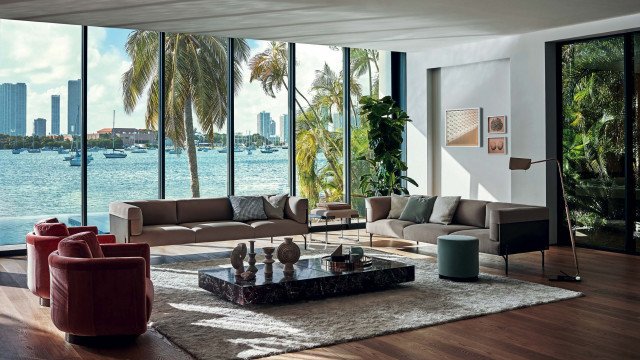 This picture shows a luxurious and modern lounge room with stylish furnishings. The room has a light gray sofa, two black armchairs, a dark wood coffee table, and an ornate floor lamp. The walls are painted in a light blue color, and the room is accented with contemporary art pieces in white frames. There is also a large window which overlooks a cityscape.