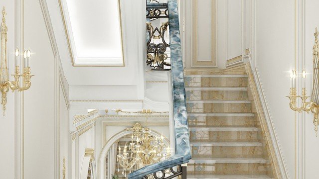jpgThe picture shows a modern interior design with a large marble staircase in the middle of an entrance hall. The walls are filled with intricate carvings, there is a large grandiose mirror, and the walls and furniture are covered in luxurious gold accents. There are two marble benches on either side of the staircases, with small accent tables as well. The flooring is also marble, with a beautiful intricate design.