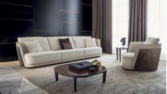 This picture is of a modern living room design. The room has luxe white furniture, an ornate area rug, and plush white sofas with a marble coffee table. There is a sleek white console table against the wall and several pieces of wall art, as well as floor to ceiling windows that provide plenty of natural light. There are also several plants scattered throughout the room for added warmth and texture.