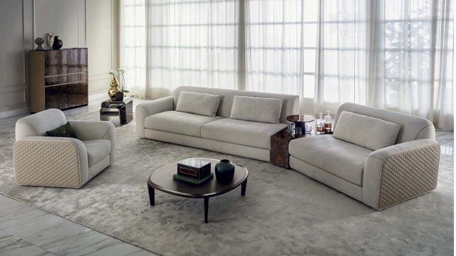 This picture is of a luxurious, spacious living room. The walls are painted a light grey and the furniture displays luxurious fabrics. The room includes several comfortable sofas and armchairs, a couple of round coffee tables and a large built-in shelf with a fireplace. There are patterned rugs on the floor and several decorative pillows in various colors scattered across the sitting area. On one side of the room, a set of French doors open to a terrace and provide plenty of natural light. The bright and airy atmosphere of this living room creates an inviting and sophisticated space.