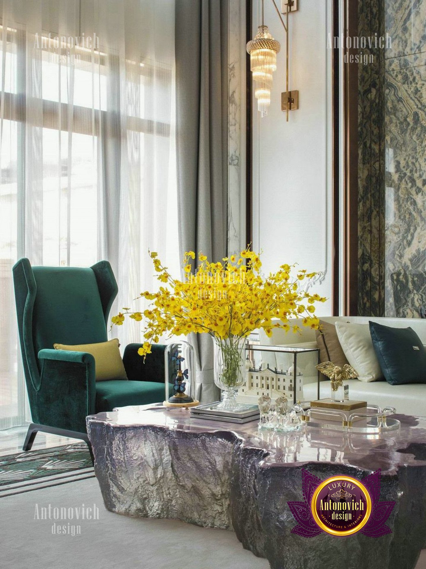 This picture shows a luxury living room with an ornate ceiling design and accents. The walls are a light shade of tan, with a white molding along the edges and a large window that overlooks a courtyard. The floor is tiled in a white marble pattern and a beige rug covers the center of the room. There is a white couch with a pink pillowed back and gold accents. In front of it is a marble-topped coffee table with two matching side tables. The walls are lined with large paintings and a few accessories. A crystal chandelier hangs from the