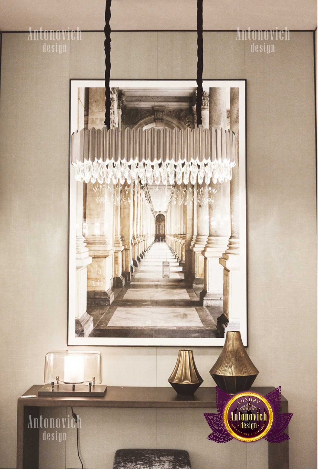 This picture shows a grand entrance hallway in an affluent home. The walls are covered with an intricate pattern of ivory and light beige tiles, while the ceiling is embellished with an ornate chandelier that gives off some soft warm light. The floor is made of dark wooden parquet panels and is offset by two area rugs. Two white stone sculptures stand out against the walls, and a flight of marble steps leads upwards towards the rest of the house.