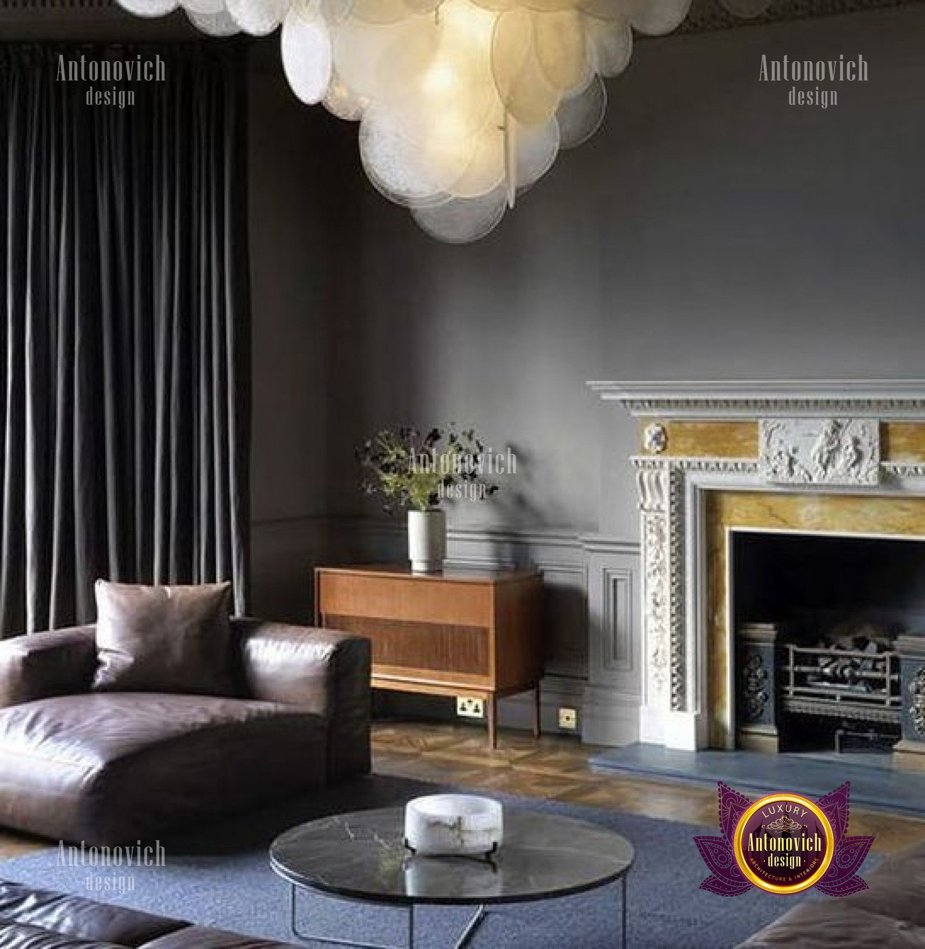 This picture shows an elegant living room interior with a gray and white color palette. The room features a luxurious velvet tufted sofa, two armchairs, an intricately carved wooden accent table, and a Persian-style rug. There are tall white curtains hanging from the windows, as well as ornate wall sconces that provide soft lighting. A few pieces of art, ranging from classic to modern, decorate the walls.