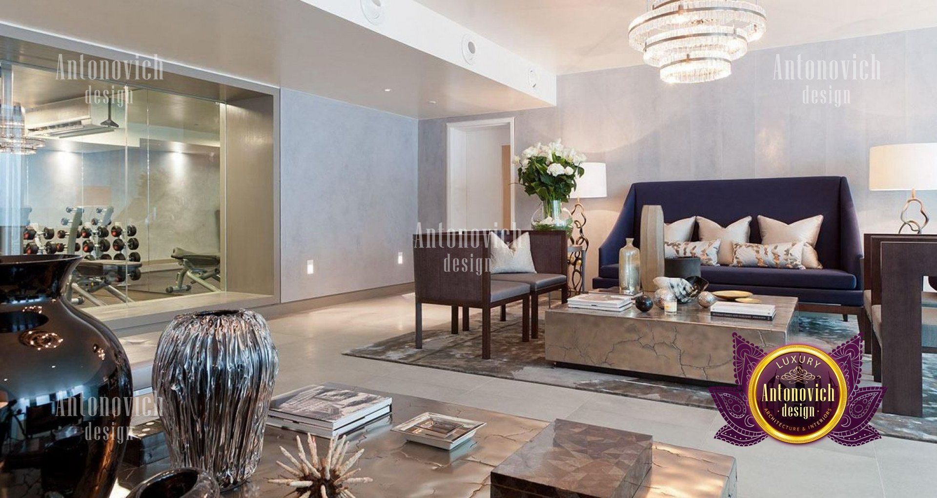 This picture shows the interior of a luxurious living room designed by Antonovich Design. The room features a polished marble floor, a large chandelier suspended from the ceiling, a grand fireplace with intricate stone details, and elegant white upholstered furniture around the room. The walls feature a soft blush pink paint finish and wide windows to let in plenty of natural light.