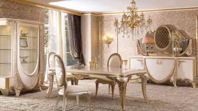 This picture shows a luxurious, elegant interior design. The interior features a marble floor, intricate metal railings, and white walls adorned with artistic and colorful wallpaper. There is an ornate white decorative ceiling and large window with heavy drapes. A marble-topped table with intricate designs sits in the center of the room and is lit by an elegant crystal chandelier. The room also includes several plush velvet couches and chairs upholstered in a rich burgundy and black detail. Several pieces of art adorn the space, creating an inviting and sophisticated atmosphere.
