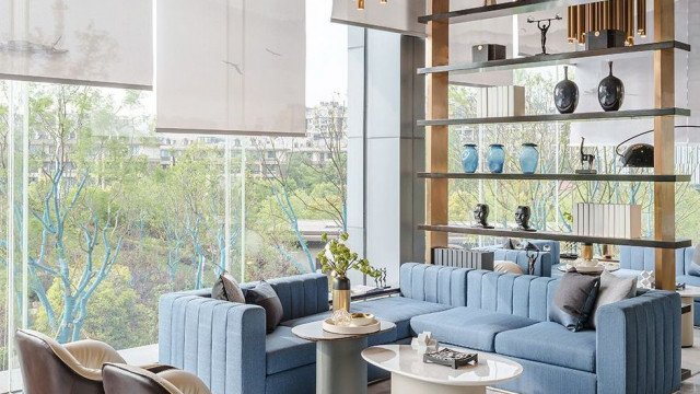 The picture is of a luxurious modern living room. It features a white sofa and chair set, with a grey accent wall in the background. The flooring is a light hardwood, and a white marble area rug is featured in front of the sofa. On either side of the sofa are glass and metal end tables with white lamps on them. Also in the room are two tall bookshelves filled with books and decor, as well as a few abstract art pieces on the walls.