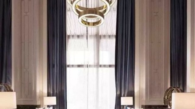 The picture shows an elegant master bedroom, featuring a traditional style and a neutral color palette. The room is decorated with an oak wood headboard, a white linen upholstered bed, an antique side table, and a pair of crystal chandeliers. The walls are painted in a light beige color and are adorned with two large landscape paintings. The flooring is a beige marble, and the window treatments are a unique combination of patterned golden curtains and sheer white drapes.