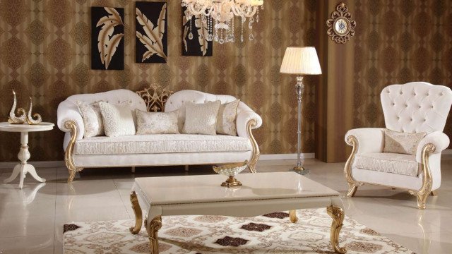 This picture shows an interior design for a luxurious living room featuring a curved sofa with a black velvet fabric and a large ornate gold coffee table in the center. The walls are a light shade of pink with gold accents, and the floor is made up of intricately patterned tiles. There is also a striking crystal chandelier hung from the ceiling and shelves of books flanking the sofa.