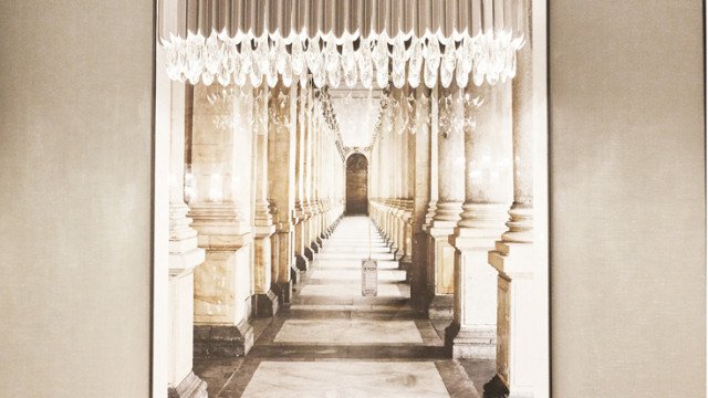 This picture shows a grand entrance hallway in an affluent home. The walls are covered with an intricate pattern of ivory and light beige tiles, while the ceiling is embellished with an ornate chandelier that gives off some soft warm light. The floor is made of dark wooden parquet panels and is offset by two area rugs. Two white stone sculptures stand out against the walls, and a flight of marble steps leads upwards towards the rest of the house.