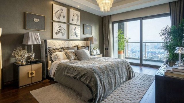 This is a picture of an elegant luxury bedroom in a modern home. The walls are painted a light beige color, with wide white trim framing the perimeter of the room. A large floor-to-ceiling window is off to one side, letting in a lot of natural light, while the center of the room features a large bed with a dark gray tufted headboard flanked by two mirrored nightstands. On the left side of the bed is a chaise lounge with a decorative pillow, while on the right is a plush white armchair and ottoman. The