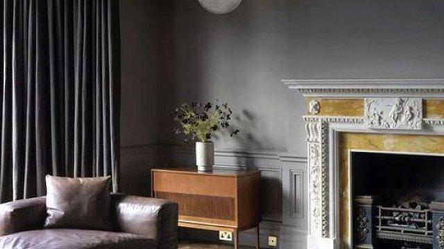 This picture shows an elegant living room interior with a gray and white color palette. The room features a luxurious velvet tufted sofa, two armchairs, an intricately carved wooden accent table, and a Persian-style rug. There are tall white curtains hanging from the windows, as well as ornate wall sconces that provide soft lighting. A few pieces of art, ranging from classic to modern, decorate the walls.