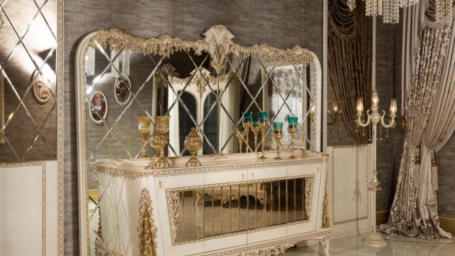 This picture shows an elegant bedroom designed by Antonovich Design, a luxury interior design company based in the UAE. The bedroom features a beautiful four-poster bed with drapes, a chest of drawers inlaid with mother of pearl, chandelier lighting, and a wooden vanity table with a chair. The colour palette of the room is neutral, with grey, white and silver accents throughout.