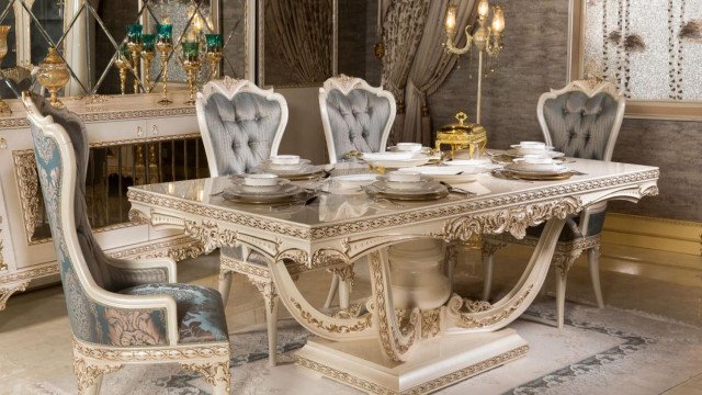 This picture shows a luxurious bedroom with gold walls and a large chandelier hanging from the ceiling. The furniture consists of an ornate four-poster bed with a white canopy, a large vanity desk with a large mirror, and two matching chairs upholstered in white fabric. There is a large rug in the center of the room and the floor is tiled with marble. On either side of the bed are small end tables.