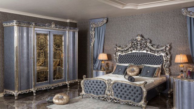 This picture shows a luxurious bedroom designed with an elegant crystal chandelier hanging from the high ceiling. The walls are adorned with beautiful wallpaper featuring exquisite patterns with gold accents. The focal point of the room is a large four-poster bed with tufted headboard and footboard. The bed is dressed in white bedding and several plush pillows. A stylish armchair is situated near the bed and a bench is positioned at the foot of the bed. To finish off the look, there is a luxurious rug laying on the floor.