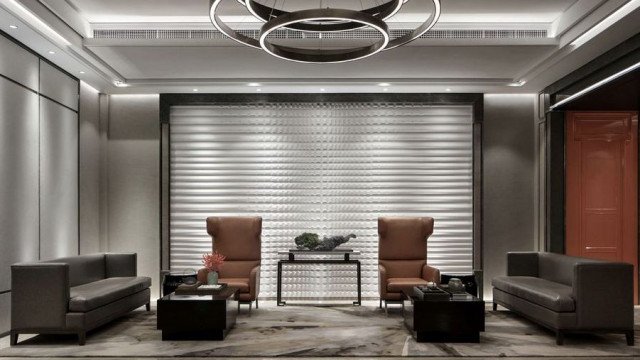 This picture shows a luxurious modern living room with tall ceilings and large windows. The walls are painted in a neutral shade of white, and the floor has a grey-toned marble tile. There is a black leather sectional sofa with decorative throw pillows in earth tones as well as a round glass coffee table. On the opposite wall is a large floor-to-ceiling stone fireplace with built-in shelving on either side. An ornate metal chandelier hangs above the sofa, and there is a painting on the wall behind it.