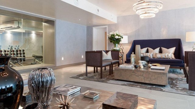 This picture shows the interior of a luxurious living room designed by Antonovich Design. The room features a polished marble floor, a large chandelier suspended from the ceiling, a grand fireplace with intricate stone details, and elegant white upholstered furniture around the room. The walls feature a soft blush pink paint finish and wide windows to let in plenty of natural light.