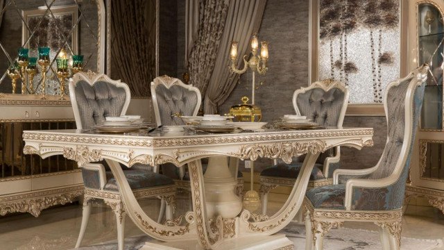 This picture shows a luxurious dining room with ornate decor. The walls are painted a deep green and have detailed wood molding around the entrance. In the center of the room is a large table surrounded by 8 chairs upholstered in velvet. On the walls hang several chandeliers and sconces, giving the room a warm and inviting feel. The finishing touches are provided by a large painting in an ornate gilded frame, and a grand piano in the background.