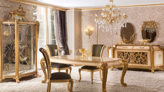 This picture shows an ornately decorated modern dining room. The walls are lined with gold and white striped wallpaper, and the ceiling has white crown molding and a striking crystal chandelier. The floor is covered in a large beige and cream-colored rug that is accented with matching furniture pieces, including a table, chairs, and sideboard.