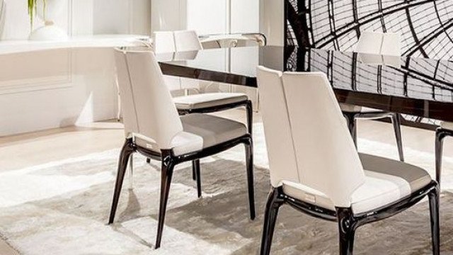 This picture shows a modern interior design featuring a black and white color palette. In the foreground, there is a large sofa with a plush grey rug beneath it, and two white armchairs on either side. There is also a white coffee table in the center of the room and a black dining table with chairs on the left. On the right, there are built-in cabinets with a variety of decorations on top, including a vase of white flowers and a ceramic pot. On the wall behind the furniture, there is a large framed painting with an abstract pattern in shades of grey and white