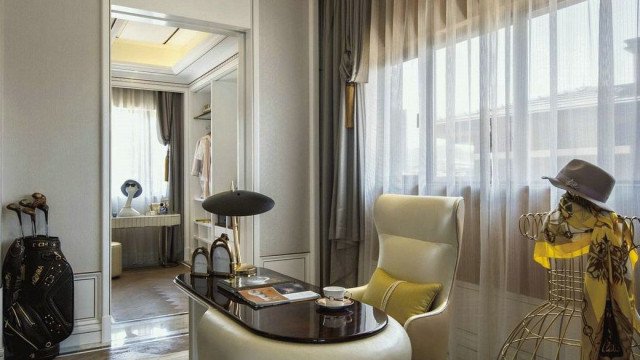 This is a picture of a modern, luxurious living room with mostly white and grey tones. The walls are adorned with light grey wallpaper with silver accents and the floor is covered with a white area rug. There is a long, sleek white sofa with several decorative cushions, and a beige velvet armchair in the corner. On either side of the sofa are two wooden tables with glass tops. A large painting of a peaceful cityscape hangs on the wall, and several framed photographs are scattered around the room. The finishing touches are delicate metal sculptures and several potted plants.