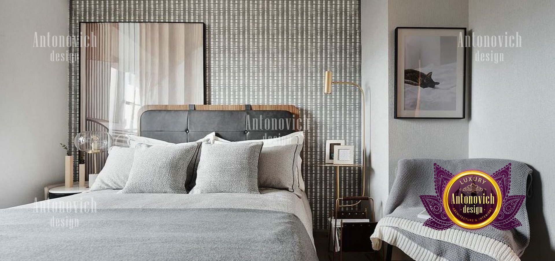 This picture shows a luxurious bedroom decorated with gold and white tones. The bed is upholstered with a gold and white striped fabric and has several matching throw pillows. On the sides of the bed are two nightstands with contemporary lamps and matching frames. The room also has a high-backed armchair, two area rugs and an ornate chandelier hanging from the ceiling. The walls are painted in a pale gray color and have several pieces of art hung over the bed.