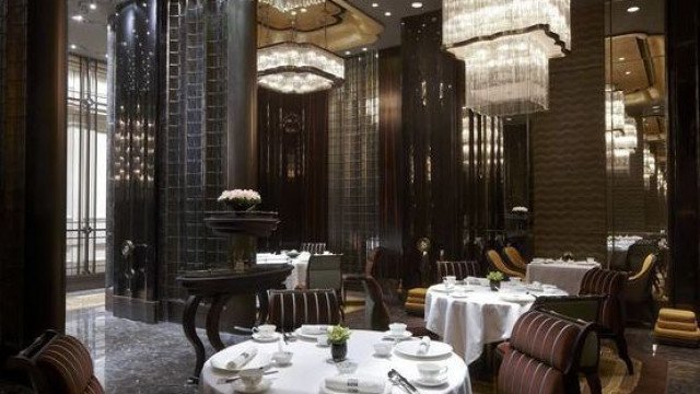 This is a picture of a luxurious, modern-style living room designed by Antonovich Design. The room features a large white leather sofa, two glass side tables with marble tops and two white leather armchairs. The walls are decorated with framed art pieces and a large flat-screen TV mounted on the wall. The floor is tiled with dark wood and the ceiling features a modern chandelier.