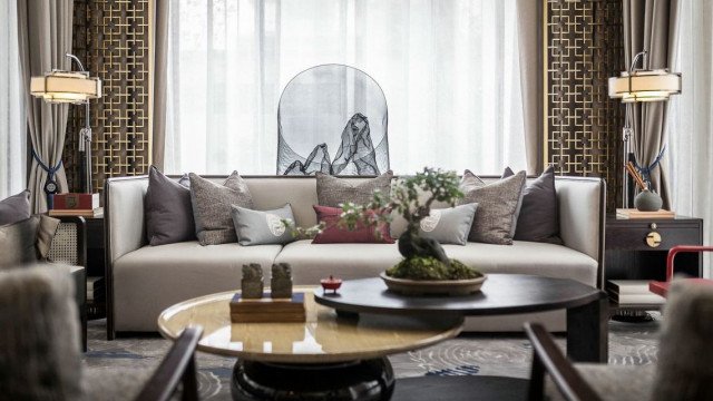 This picture shows a modern living room with an angled ceiling. The walls and ceiling are painted in light beige, while the floor is covered in dark wood. The room features an L-shaped grey sofa, with two armchairs placed opposite it. A coffee table is situated in front of the sofa and there is an abstract painting on the wall behind it. The ceiling has several recessed lights, as well as a chandelier.