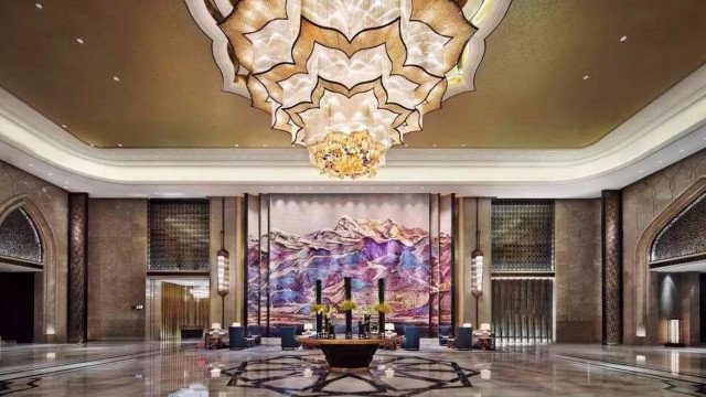 The picture shows a luxurious hallway with a unique ceiling design, a gold chandelier and walls with crystal patterns. The floor is made of white marble and the walls are painted in a dark grey hue. The hallway is illuminated by a row of small lamps mounted on the wall. At the end of the hallway, there is a doorway with a large glass window and a staircase leading to the upper levels of the building.