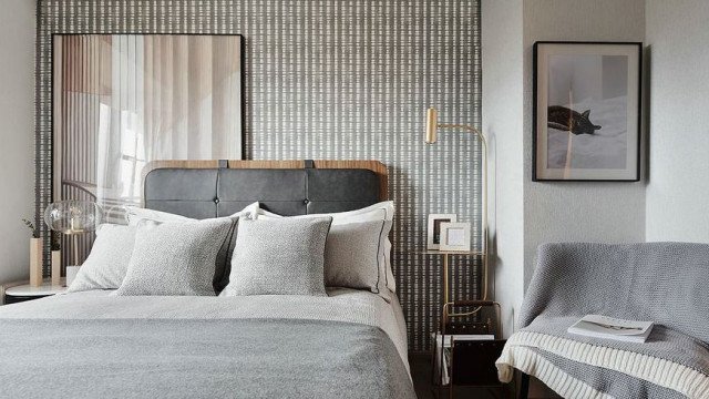 This picture shows a luxurious bedroom decorated with gold and white tones. The bed is upholstered with a gold and white striped fabric and has several matching throw pillows. On the sides of the bed are two nightstands with contemporary lamps and matching frames. The room also has a high-backed armchair, two area rugs and an ornate chandelier hanging from the ceiling. The walls are painted in a pale gray color and have several pieces of art hung over the bed.